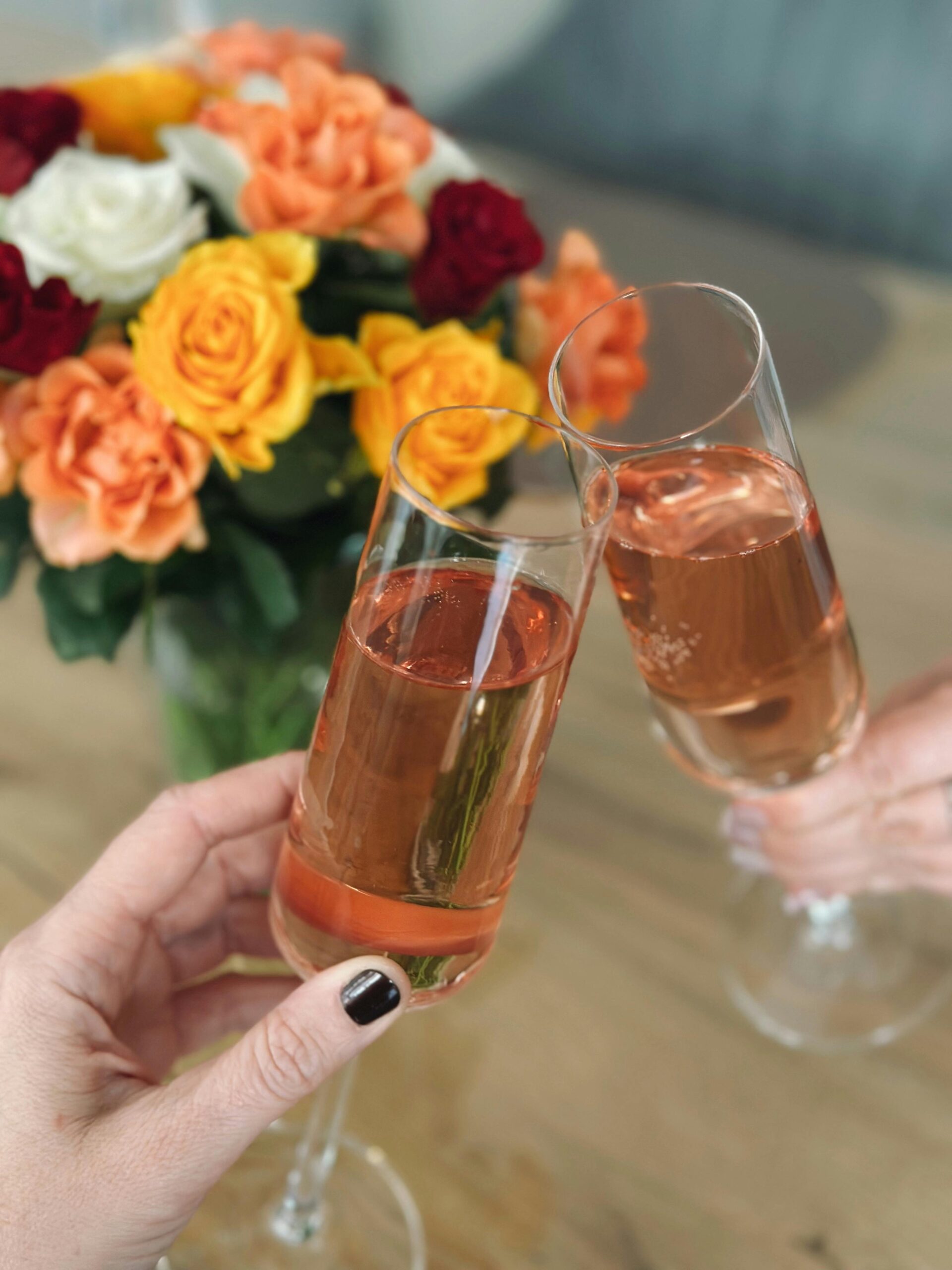 Two hands clinking champagne glasses with a bouquet of vibrant roses in the background.